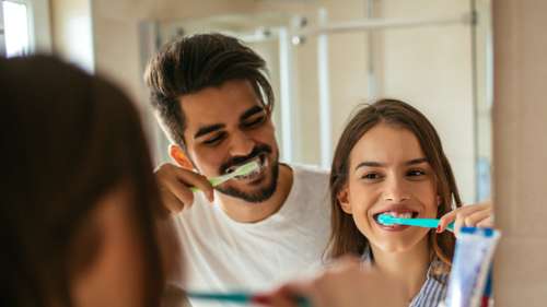 A man and woman brushing their teeth together in a bathroom mirror